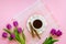 White porcelain cup with coffee, two coffee rols on saucer with wavy edge and and bouquet of pink or purple tulips on a pink table