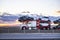 White and red classic car hauler semi truck transporting cars on the hydraulic semi trailer driving on the road at twilight