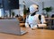A white robot sits in a cafe with a laptop. AI generated