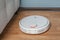 White Robot vacuum cleaner runs in corner near sofa on wood parquet floor. Modern smart cleaning technology housekeeping