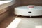 A white robot vacuum cleaner vacuums along the skirting Board near a large window through which the bright sun shines. close-up,