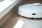 A white robot vacuum cleaner vacuums along the skirting Board near a large window through which the bright sun shines. close-up,