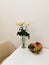 White roses in a glass vase, on a white table with a fruit plate with grapes, avocados, persimmons, mangoes and apples