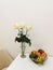 White roses in a glass vase, on a white table with a fruit plate with grapes, avocados, persimmons, mangoes and apples