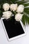 White roses and petals lying on a white surface. tablet with empty screen. Styled mock up top view on pale peach background