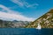 A white sailboat floats on the waters of the Bay of Kotor against the backdrop of mountains and blue sky.