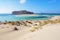 The white sand beside the sea, where the bottom can be seen, the beach with people. Beautiful mountains on a blue horizon.