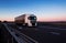 A white semi-trailer truck drives down the highway in the evening with its headlights on. Blue sky and sunset. The mode