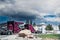 White semi truck pulls two new red semis into fuel service station under stormy skies