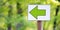 White sign or label with green arrow on stick on path in green spring or summer forest. Notification or direction of movement,