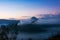 White smoke coming out of volcanoes surrounded by white clouds of mist and a clear blue sky.