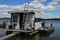 White square shaped house boat with ladder leaning on front wall and wooden bridge in front