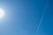 A white streak from an airplane in a summer blue sky with a circle of bright sun.