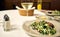 A white table at a restaurant with a glass of wine, appetizer plate of bacon and salad, and bottle of salt and pepper