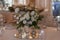 White tablecloths with clear vases and white flowers and fern arrangements. Golden colored plates, peavh napkins, table