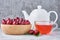 White teapot, herbal tea in glass cup and fresh cherry in wooden bowl on a gray background