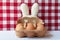White toweling bunny ears with a carboard mask and eggs. White and red checkered background and white lace tablecloth