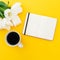 White tulips, notebook with mug of coffee on yellow background. Flat lay, top view.