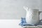 White Vintage Ceramic Cream Milk Jug Standing on Folded White and Blue Cotton Linen Napkins on Table. Holiday Baking Workshop