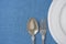 White Vintage Empty Plate Half Circle Silver Fork Spoon on Light Blue Linen Fabric. Minimalist Japanese Style Table Setting