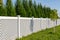 White vinyl fence in a cottage village. Several panels are connected by columns. Tall Thuja bushes behind the fence. Fencing of