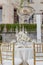 White wedding table with luxurious setting, bouquet of roses, under crystal chandelier on terrace.