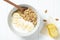 white wheat and granola with low-fat yogurt in a white bowl in a composition with a spoon, honeycombs, banana on white wooden