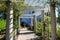 A white wooden awning in the garden covered with lush green plants and colorful flowers with stone pillars and a water fountain