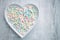 White wooden plate heart shaped filled with colourfull pastel mini marshmallows flaylay on white grey wooden underground