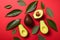 Whole and cut ripe avocadoes with green leaves on red background, flat lay. Minimalistic concept. Avocado assortment background,