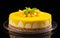 Whole mango cheesecake topped with yellow glaze and pieces of mango with a mint leaf on a plate against black background