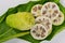 Whole Noni fruit Morinda citrifolia and slices cut crosswise, on leaf, against white background