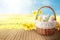 Wicker basket with festively decorated Easter eggs and daffodils on wooden table on sunlit green meadow, space for text