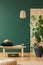 Wicker chandelier above wooden bench with emerald green pillow and hat, copy space on the empty green wall