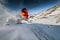 Wide angle male skier in orange suit makes a jump from a snowy ledge in the mountains. Snow powder trailing behind the