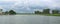 A wide angle shot of a beautiful landscape with a water pond in the middle, crop plantations around and cloud covered sky above.