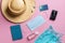 Wide brimmed straw hat, passport cover, medical face mask, hand sanitizer, credit card and small camera on a pink background.