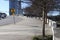 A wide curved sidewalk near the street lined with bare winter trees along the sidewalk and a metal fencing and cars driving