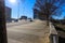 A wide curved sidewalk near the street lined with bare winter trees along the sidewalk and a metal fencing and cars driving