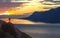 Wide mountain lake panorama. Small silhouette of tourist with backpack on rocky mountain slope with raised hands on lake water