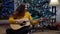 Wide shot of confident cheerful Middle Eastern young woman playing guitar singing sitting at decorated Christmas tree on