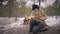 Wide shot focused bearded old indigenous man sitting in winter forest with fire burning at background. Concentrated