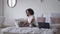 Wide shot of focused beautiful African American woman sitting on bed typing on laptop keyboard. Portrait of concentrated
