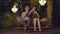 Wide shot of happy loving couple sitting on romantic retro bench with garland lights at the background and talking