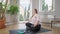 Wide shot of happy plump young woman turning on calm music in headphones and meditating sitting on exercise mat at home