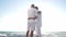 Wide shot of hugging Asian man and senior woman standing in sunshine on beach with turquoise foamy Mediterranean waves