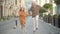 Wide shot of joyful grandfather and granddaughter holding hands and jumping as strolling on sunny city street. Portrait