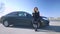 Wide shot portrait of confident female auto racer standing at black car on empty road outdoors on sunny spring or autumn