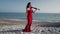 Wide shot portrait of slim beautiful Caucasian woman in red dress standing in sunshine on ocean coast with violin and