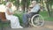 Wide shot of senior Caucasian woman in white dress sitting on bench in summer park and talking to sad paralyzed man in